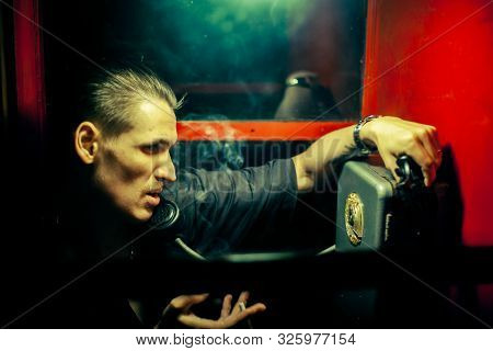 A Man Of Criminal Appearance In A Payphone Booth At Night. Talking On The Phone With A Revolver And