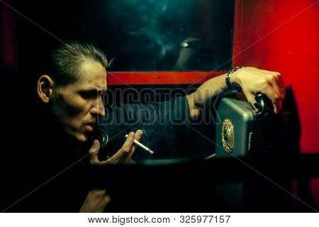A Man Of Criminal Appearance In A Payphone Booth At Night. Talking On The Phone With A Revolver And