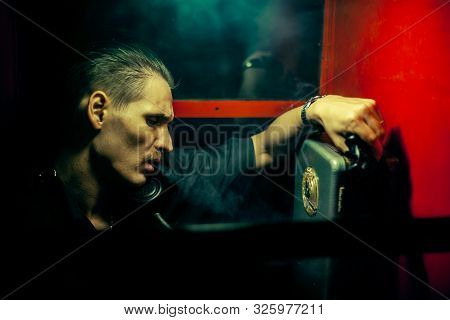 A Man Of Criminal Appearance In A Payphone Booth At Night. Talking On The Phone With A Revolver And