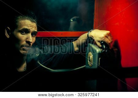 A Man Of Criminal Appearance In A Payphone Booth At Night. Talking On The Phone With A Revolver And