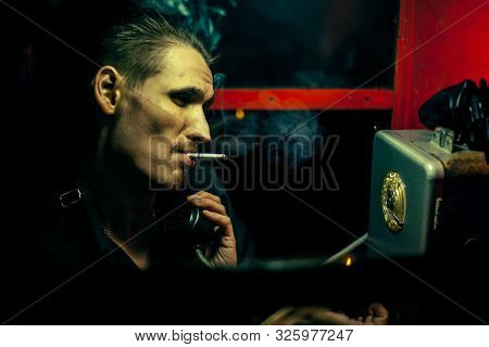 A Man Of Criminal Appearance In A Payphone Booth At Night. Talking On The Phone With A Revolver And