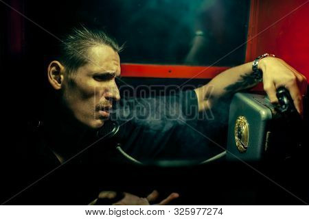 A Man Of Criminal Appearance In A Payphone Booth At Night. Talking On The Phone With A Revolver And