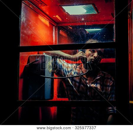 A Man Of Criminal Appearance In A Payphone Booth At Night. Talking On The Phone With A Revolver And