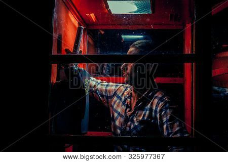 A Man Of Criminal Appearance In A Payphone Booth At Night. Talking On The Phone With A Revolver And