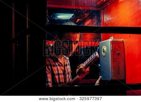 A Man Of Criminal Appearance In A Payphone Booth At Night. Talking On The Phone With A Revolver And