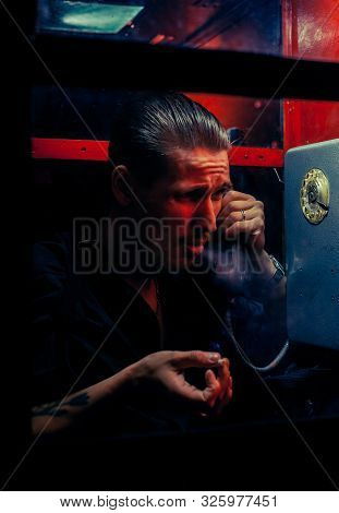 A Man Of Criminal Appearance In A Payphone Booth At Night. Talking On The Phone With A Revolver And
