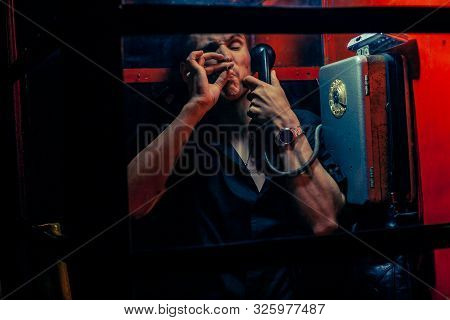 A Man Of Criminal Appearance In A Payphone Booth At Night. Talking On The Phone With A Revolver And