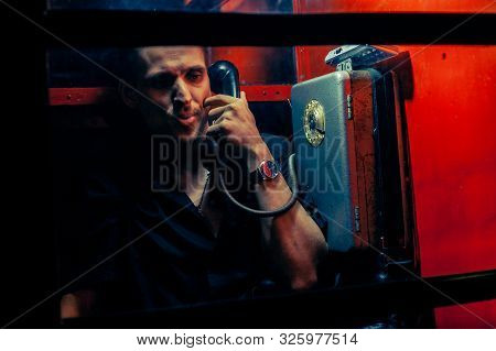 A Man Of Criminal Appearance In A Payphone Booth At Night. Talking On The Phone With A Revolver And