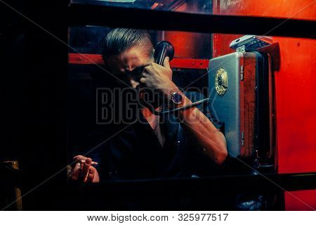 A Man Of Criminal Appearance In A Payphone Booth At Night. Talking On The Phone With A Revolver And