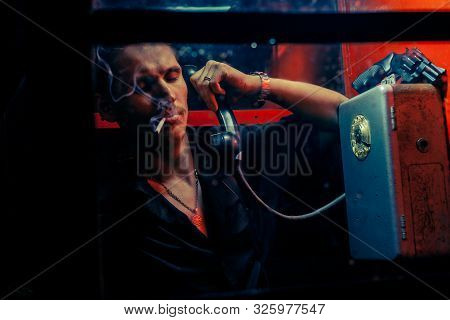 A Man Of Criminal Appearance In A Payphone Booth At Night. Talking On The Phone With A Revolver And