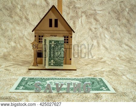 A Piggy Bank With A Dollar Stands On A Dollar Near A Wooden House.