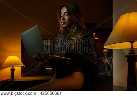 Beautiful girl in a kombidres sits on a dresser with a laptop and a skeleton in a cupboard. Many people cannot part with their ghosts.