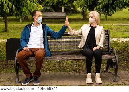 People Sitting On Park Bench In The Sun Practicing Social Distancing In Corona Crisis Wearing Masks