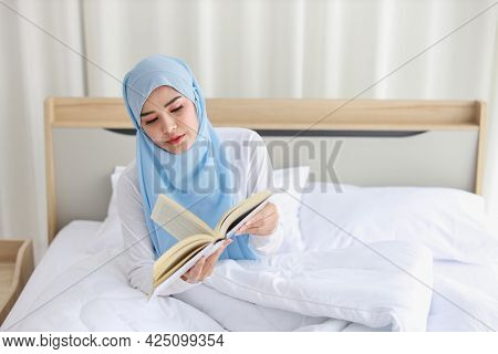 Happy Young Beautiful Asian Muslim Woman In Sleepwear Sitting, Reading Book On Bed. Cheerful And Cut