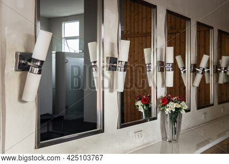 White Luminous Lamps Are Arranged In A Row Vertically On White Wall Near The Bathroom Large Mirror. 