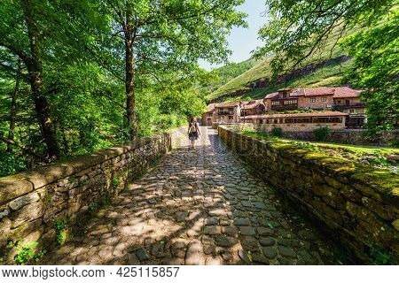 Woman Passing Over An Old Stone Bridge Towards The Village Of Old Houses. Barcena Mayor Santander.