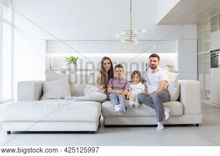 Family Portrait Of Young Parents With Their Two Children At Home, White Modern Desgin Of Living Room