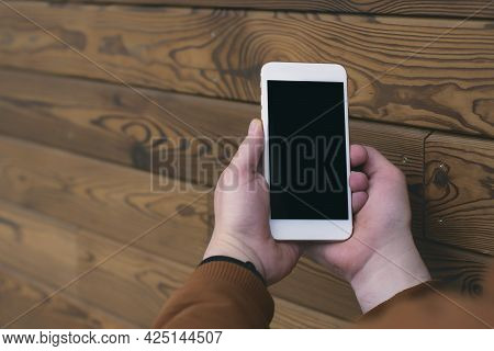 Man Holds A Mock-up Of A Smartphone With A Black Screen In His Hands On A Wooden Background.