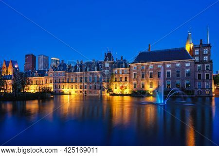 View of the Binnenhof House of Parliament and the Hofvijver lake with downtown skyscrapers in background illuminated in the evening. The Hague, Netherlands