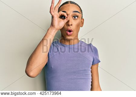Hispanic man wearing make up and long hair wearing casual t shirt doing ok gesture shocked with surprised face, eye looking through fingers. unbelieving expression. 