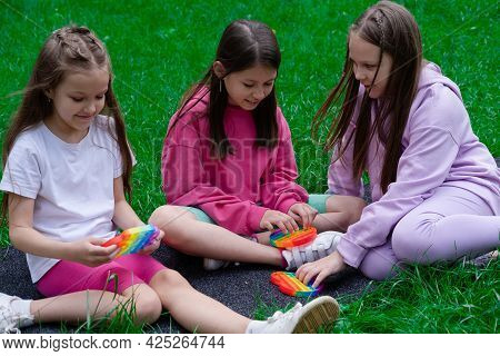 Three Teenage Girls In Colorful Clothes Play With Trendy Sensory Toy Pop It Outdors. Rainbow Soft Si