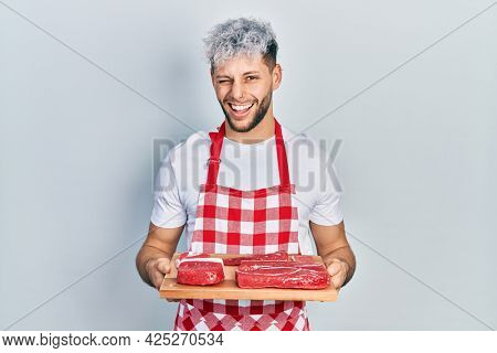 Young hispanic man with modern dyed hair holding board with raw meat winking looking at the camera with sexy expression, cheerful and happy face. 