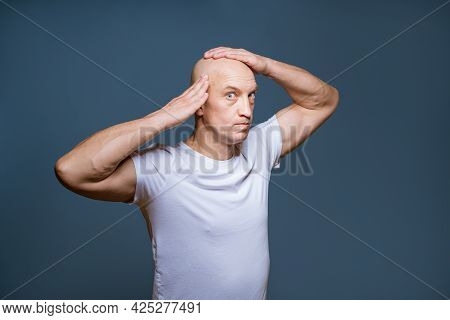 Funny Bald Man Posing On A Blue Background With Excited Facial Expressions And Raised Hands At His H