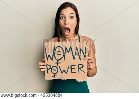 Young latin woman holding woman power banner afraid and shocked with surprise and amazed expression, fear and excited face. 