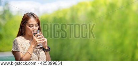 Asian Woman Drinks Water From Tall Glass Of Water At Outdoor Tree Bokeh Green For Banner Background.