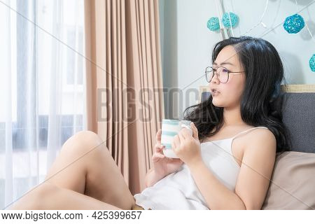 Asian Glasses Woman Holds A Cup Of Coffee And Relax On The Bed In The Morning At Her Bedroom.