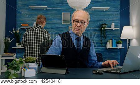 Elderly Man Using Tablet And Laptop In Same Time Compering Financial Graphs Working From Home Sittin
