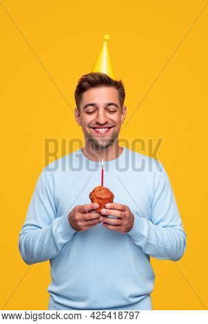 Smiling Dreamy Young Man In Party Hat Holding Festive Muffin With Candle And Making Wish With Eyes C
