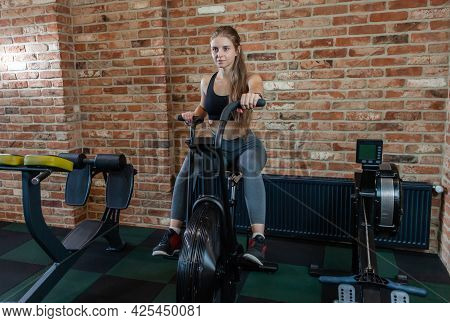Attractive Woman Using Exercise Bike At Gym. Fitness Female Using Air Bike For Cardio Workout At Fit