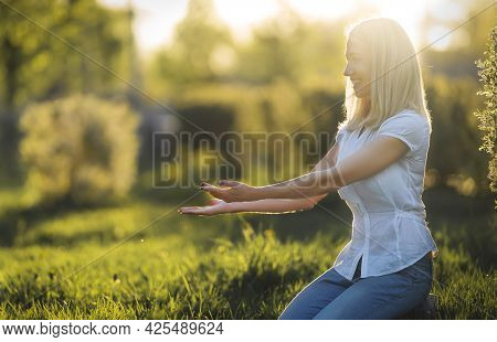 Woman With Blonde Hair Stretches Out Her Hands To Someone Tto, Sitting On The Grass At Sunset