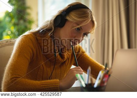 Caucasian woman working in living room at home, wearing headphones and using laptop, concentrating. flexible working from home with technology.