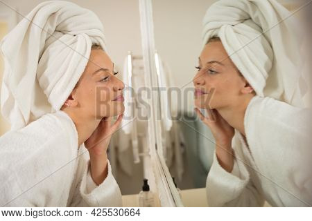 Smiling caucasian woman in bathroom with towel on head, looking in mirror and moisturising face. health, beauty and wellbeing, spending quality time at home.