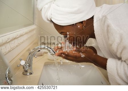 African american woman in bathroom with towel on head, standing at basin washing face. health, beauty and wellbeing, spending quality time at home.