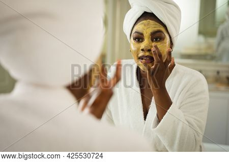 African american woman in bathroom wearing towel on head, looking in mirror and applying face mask. health, beauty and wellbeing, spending quality time at home.