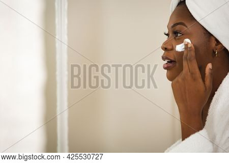 African american woman in bathroom with towel on head, looking in mirror moisturising face. health, beauty and wellbeing, spending quality time at home.