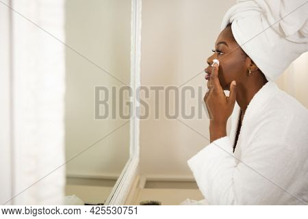 African american woman in bathroom with towel on head, looking in mirror moisturising face. health, beauty and wellbeing, spending quality time at home.