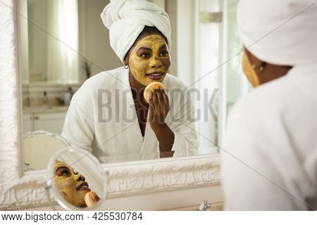 Happy african american woman in bathroom with towel on head, looking in mirror applying face mask. health, beauty and wellbeing, spending quality time at home.