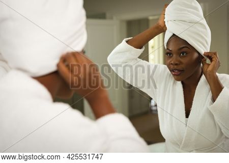 African american woman in bathroom wearing bathrobe, looking in mirror putting towel on head. health, beauty and wellbeing, spending quality time at home.