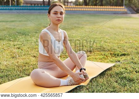 Peaceful Sporty Woman Sitting On Karemat In The Lotus Pose In Stadium, Wearing White Top And Beige L