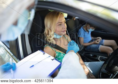 Medical worker wearing ppe suit taking swab tests of caucasian mother with son sitting in car. medical and health precautions, lifestyle during covid 19 pandemic.
