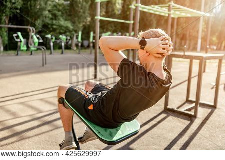 Strong sportive man guy pumps his abs at the stadium outdoors. Adult male person during workout for tummy with additional sport equipmant. Sportsman exercising outside