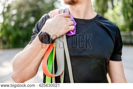 Sportive man guy holding in his hand wearing fitness tracker set of colorful elastic rubber bands at the stadium outdoors. Athlete male person during workout with additional sport equipmant