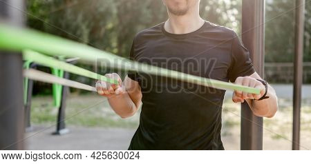 Strong man guy training with elastic rubber band at the stadium outdoors. Adult male person during workout for arms and chest with additional sport equipmant. Sportsman exercising outside