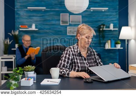 Positive Senior Freelancer Opening Laptop Sitting At Home Office And Mature Husband Reading A Book. 