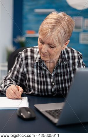 Mature Entrepreneur Taking Notes On Notebook Working In Home Office. Elderly Woman In Home Living Ro