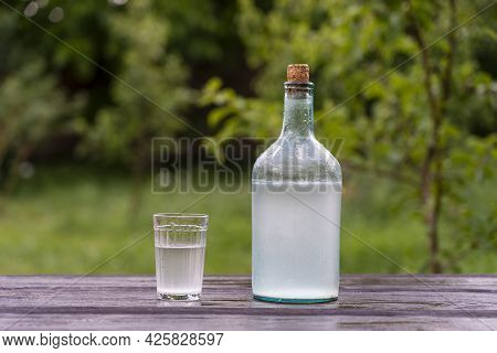 Big Bottle And Glass Of Moonshine Or Vodka On Wooden Table In Sunny Summer Day In Garden, Close Up